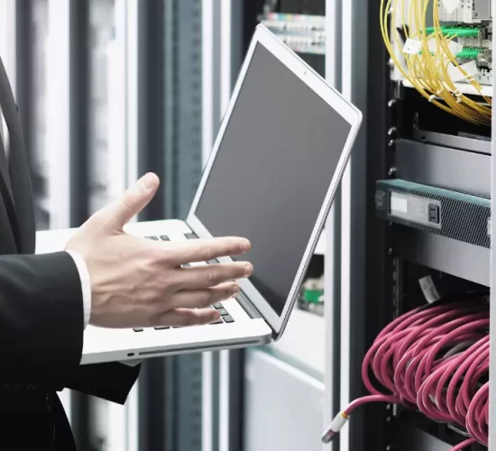 Vista de la mano de un hombre sujetando una laptop frente al cableado de un servidor